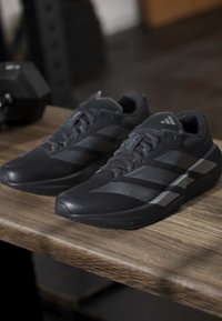 Ζευγάρι μαύρα παπούτσια για τρέξιμο Adidas με γκρι ρίγες τοποθετημένα σε ξύλινο πάγκο δίπλα σε αλτήρα σε γυμναστήριο.
