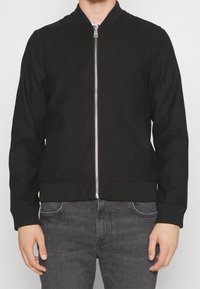 Black bomber jacket with a smooth fabric, ribbed cuffs and hem, front zipper, and two side pockets. Paired with gray denim pants.