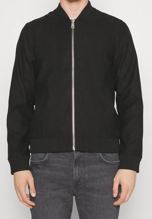 Black bomber jacket with a smooth fabric, ribbed cuffs and hem, front zipper, and two side pockets. Paired with gray denim pants.