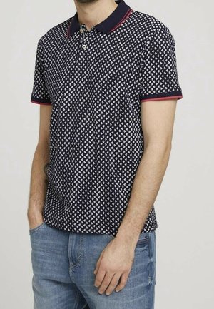 Homme portant un polo bleu marine avec motif blanc, bordure rouge au col et aux manches, assorti à un jean bleu clair, mains dans les poches.
