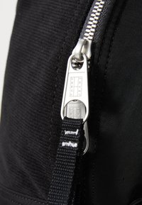Juodas audinio kuprinės, pasižyminčios sidabriniu metaliniu užtrauktuku, tekstūruotu paviršiumi ir juoda juosta su baltu tekstu. Etiketėje užrašyta "TOMMY JEANS."