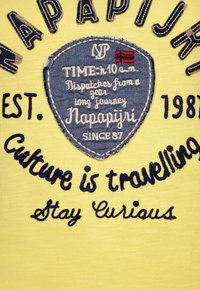 Camisa de tela amarilla con un parche de logo de denim azul y texto bordado, que destaca temas de viaje y curiosidad en hilo oscuro.
