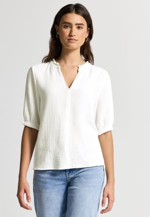 Blouse blanche à manches courtes avec un tissu texturé, décolleté en V et bordures volantes. Assortie à un jean en denim bleu clair.