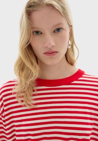 Rood-wit gestreepte gebreide top met een ribbelachtige ronde hals. Details omvatten een zachte textuur en een relaxte pasvorm. Het haar van het model is gedeeltelijk zichtbaar.