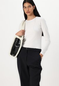 Un sac à main synthétique noir avec un bord blanc duveteux, de forme rectangulaire et avec une longue poignée. Le modèle porte une chemise blanche ajustée à manches longues et un pantalon sombre.