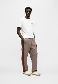 Λευκό μπλουζάκι με πράσινο κείμενο, καρό παντελόνι με καφέ ρίγα στο πλάι και μπεζ αθλητικά παπούτσια. Το μοντέλο φοράει ελαφριά γυαλιά ηλίου.