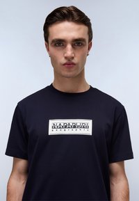 Navy blue cotton t-shirt featuring a white rectangular logo print that reads "NAPAPIURI geographic" centered on the chest.