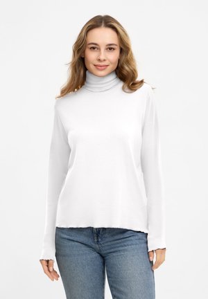 Woman with light brown wavy hair wears a white turtleneck sweater and blue jeans against a plain white background.