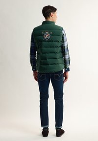 Chaleco acolchado verde con acolchado horizontal, logo bordado en la parte posterior, combinado con una camisa de cuadros, jeans oscuros y zapatos de ante marrón.