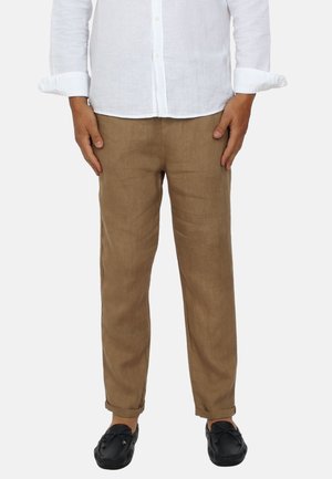Uomo che indossa pantaloni di lino beige, camicia bianca a maniche lunghe con bottoni e polsini arrotolati, e scarpe nere slip-on, in piedi davanti a uno sfondo semplice.