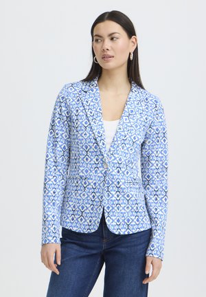 Vrouw met een blauw gebloemd colbert over een wit shirt en donkere jeans, staand met ontspannen handen, kijkend naar opzij tegen een effen achtergrond.