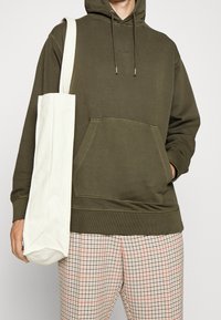 Olivgrön hoodie med dragsko, framficka och ribbad nederkant, tillsammans med rutiga byxor i beige och röd. Håller en vit canvas tote bag.