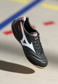 Pantofii de fotbal indoor din piele neagră cu accente albe, suprafață texturată, vârf rotund, șireturi negre și talpă din cauciuc de culoare caramel. Prezintă detalii roșii.