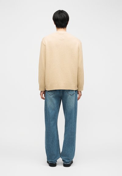 Homme aux cheveux noirs courts portant un pull beige et un jean bleu ample, debout de dos sur un fond clair uni.