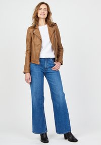Brun læder bikerjakke, hvid t-shirt med rund hals, blå vide jeans og sorte hæle støvler. Model står imod en neutral baggrund.