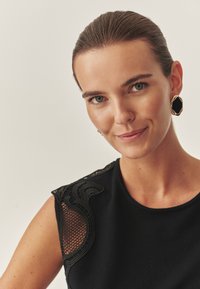 Fekete felső, hálós részletekkel a vállaknál, kerek nyakkivágással és sima textúrával. A modellen merész arany és fekete fülbevalók vannak. Világos háttér.