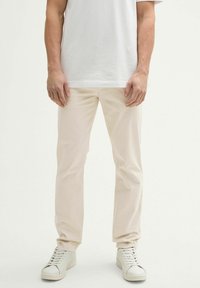 Beige chinos ar slaidu piegriezumu, izgatavoti no kokvilnas, ar gludu tekstūru. Apvienoti ar baltiem sporta apaviem. Modelis stāv taisni.