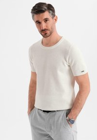 Camiseta de punto blanca de manga corta con un patrón texturizado, escote redondo y diseño ajustado, combinada con pantalones gris claro y un reloj plateado.
