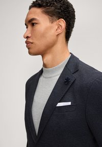 Marineblazer mit strukturierter Oberfläche, abgesteppten Revers, einem grauen Rollkragenpullover und einer marineblauen Blumenstecknadel am Revers. Verfügt über ein weißes Einstecktuch.