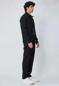 Veste en denim noir avec une fermeture à boutons, deux poches poitrine et des manches longues, associée à un pantalon noir assorti et des baskets blanches.