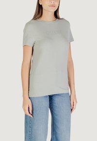 T-shirt in cotone grigio chiaro con maniche corte, con un logo "GUESS" in rilievo centrato. Abbinato a jeans in denim blu a gamba larga.