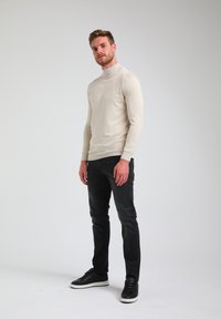 Svetlo bež pulover s turtleneck ovratnikom, prilegajoča zasnova, rebrasti manšeti, v kombinaciji s črnimi kavbojkami in temnimi supergami, na enobarvni podlagi.