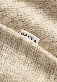 Bézs színű kötött szövet, amelyen jól látható egy fehér címke, rajta fekete betűkkel a "GABBA" felirat.