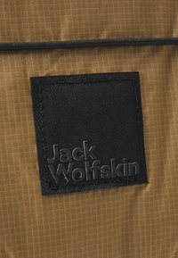 Brauner strukturierter Stoff mit einem schwarzen rechteckigen Patch. Der Patch enthält den eingeprägten Text "Jack Wolfskin" und ein dezentes Gittermuster im Hintergrund.