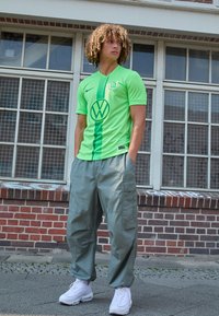 Maglietta da calcio verde con strisce verticali e logo VW, abbinata a pantaloni cargo grigi larghi e sneakers bianche, indossata da un modello all'aperto.