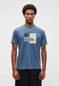 Homem com cabelo afro, vestido com uma t-shirt gráfica azul com design abstrato e calças escuras, de pé contra um fundo claro liso.