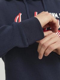 Felpa blu navale con polsini a coste, caratterizzata da testo bianco e rosso. In primo piano, una mano che aggiusta la manica, evidenziando la trama del tessuto.