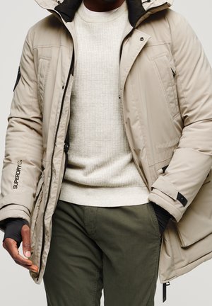 Homme portant une veste matelassée beige par-dessus un pull en maille crème et un pantalon vert, une main partiellement dans la poche de la veste, l’autre détendue.