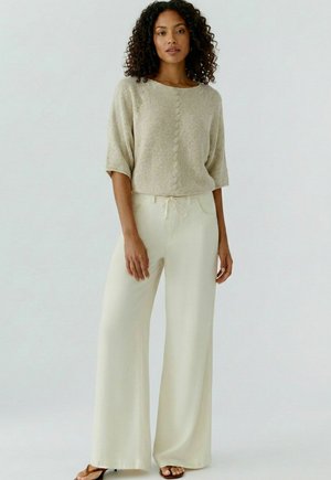 Femme aux cheveux bouclés portant un haut en maille texturée beige à manches mi-longues et un pantalon large crème avec taille à cordon.