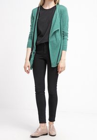 Teele cardigan med åpen front, lange ermer, sammen med en svart topp og trange svarte jeans. Lyse beige sko med glatt finish.