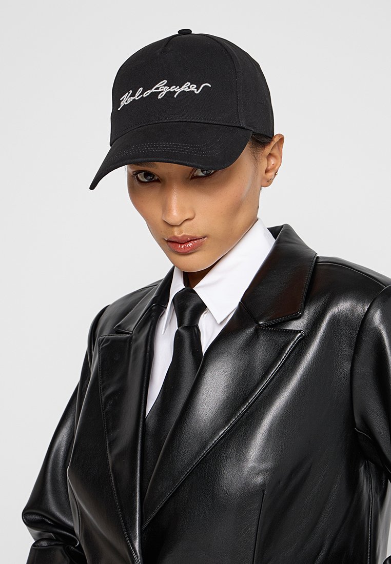 Schwarze Baseballmütze mit gesticktem Text, getragen mit einer glänzenden schwarzen Lederjacke und einem weißen Hemd, das einen eleganten Kragen und eine Krawatte aufweist.