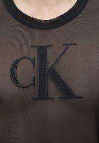 Άτομο που φοράει διάφανο μαύρο τοπ με μεγάλα μαύρα κεντημένα γράμματα «cK» στο κέντρο του στήθους.