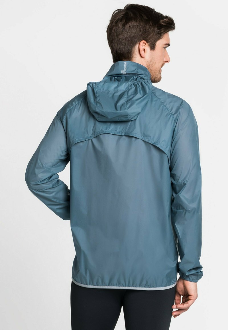 Mężczyzna odwrócony tyłem, ubrany w niebieską kurtkę z kapturem i czarne spodnie na tle jednolitej białej ściany.