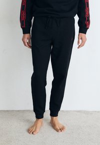 Schwarze Jogginghosen aus einem strukturierten Material, mit elastischem Bund und Kordelzug. Verfügt über ein dezentes rotes Logodetail an der Vorderseite und den Bündchen.