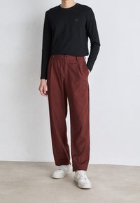 Black long-sleeve shirt with a square logo, paired with loose-fitting maroon trousers; person wears white sneakers. Smooth textures.