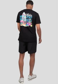 Camiseta de algodón negra con un diseño gráfico colorido en la parte posterior, que presenta plantas tropicales, flamencos y tablas de surf. Usada con pantalones cortos negros.