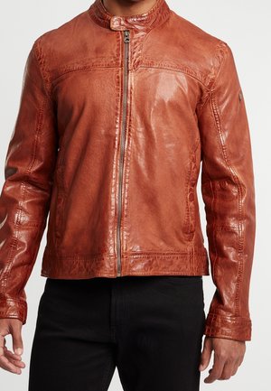Man draagt een bruine leren jas met een rits aan de voorkant, staat met ontspannen armen langs zijn zij, draagt een zwarte broek.