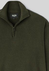 Pull-over vert foncé avec un col côtelé et une fermeture éclair, fabriqué dans un tissu doux et texturé. L'étiquette indique "BZB, taille L."