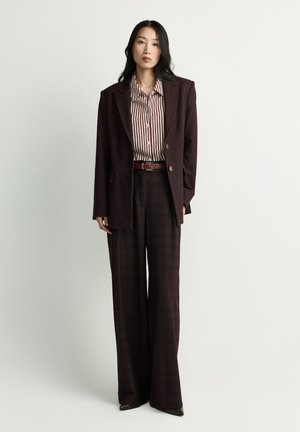 Woman standing wearing dark brown blazer and wide-leg pants, striped button-up shirt, and red belt against plain light background.