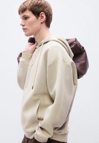 Μπεζ φούτερ με κουκούλα και ριγωτή υφή, με μπροστινή τσέπη, κορδόνι με τσόκερ και καφέ αντίθετο σακίδιο πλάτης κρεμασμένο στον ώμο.