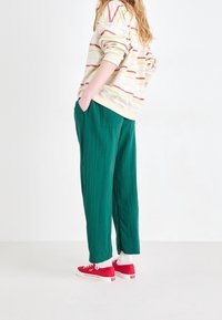 Groene geplooide broek met zijzakken, gecombineerd met een gestreepte crème sweatshirt en rode sneakers met witte zolen en sokken.