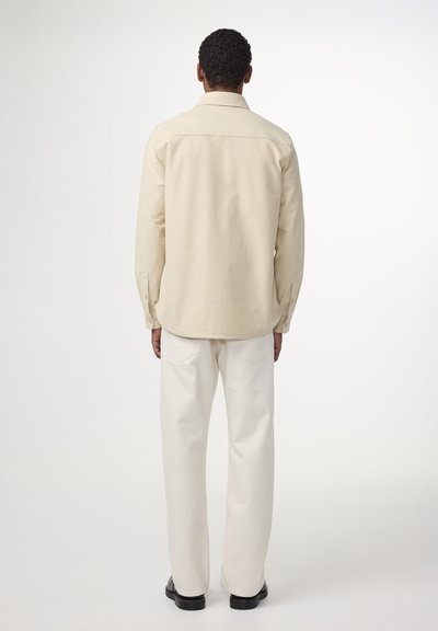 Beige skjorta med knappar och långa ärmar, som har en standardkrage och en rak fåll, ihop med off-white raka byxor.