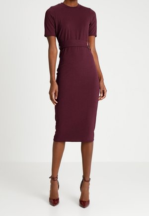 Nainen pukeutuneena istuvaan, ribbineulottuun burgundinpunaisen midi-mekkoon, jossa on lyhyet hihat ja vyötärökoriste, yhdistettynä samansävyisiin, teräväkärkisiin korkokenkiin.