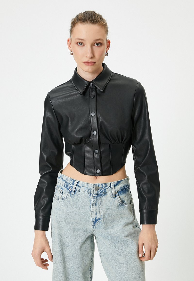 Schwarze Kunstleder-Cropped-Jacke mit Kragen, Knopfschluss und langen Ärmeln; kombiniert mit hellblauen Jeans.