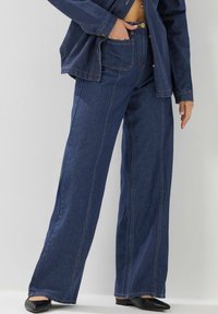 Pantalons en denim à jambes larges de couleur bleu foncé avec des coutures contrastées, présentant une taille haute, des poches avant et une texture lisse.