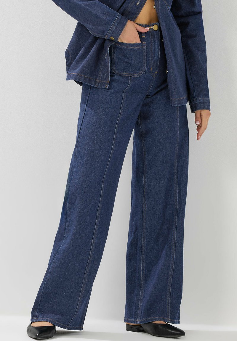 Pantalons en denim à jambes larges de couleur bleu foncé avec des coutures contrastées, présentant une taille haute, des poches avant et une texture lisse.